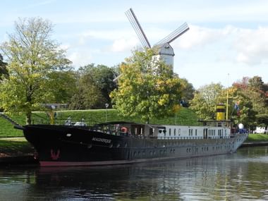 Black river cruise ship Magnifique I moored along a tree-lined canal with a white windmill visible on the green embankment behind it.