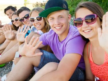 Six smiling teenagers wearing sunglasses sitting together outdoors, waving at the camera in a friendly group selfie pose.