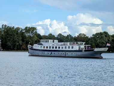 Gray and white passenger ship Floor van Aemstel anchored on calm water with trees in the background and white clouds in blue sky.