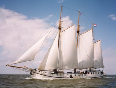 Dreimastiges Segelschiff Elizabeth unter vollen weißen Segeln auf offener See. Niederländische Flagge am Mast vor blauem Himmel mit weißen Wolken.