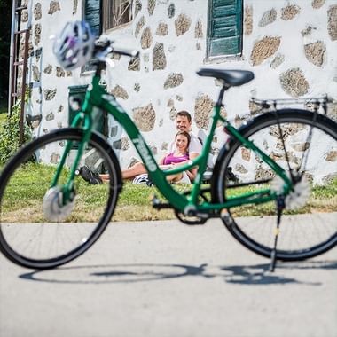 Grünes Fahrrad im Vordergrund mit zwei Radfahrern, die auf dem Rasen vor einer weißen Steinmauer mit grünen Fensterläden rasten.