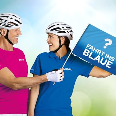 Zwei lächelnde Radfahrer mit Helmen halten eine blaue Flagge mit Text 'Fahrt ins Blaue'. Einer trägt ein pinkes, der andere ein blaues Shirt.