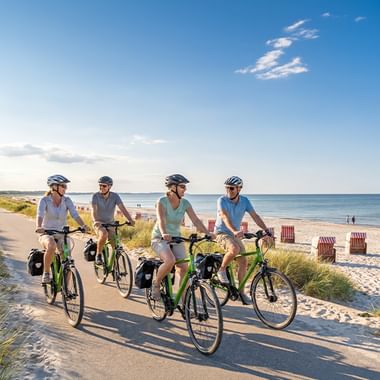 Vier Radfahrer mit Helmen und bepackten Rädern auf einem sandigen Küstenweg. Strand mit Strandkörben und ruhiger See im Hintergrund.