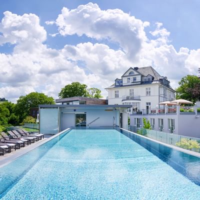 Dach-Infinity-Pool der Villa Hügel Trier mit Glasgeländer, Liegestühlen und weißem Villengebäude unter dramatischem Wolkenhimmel.