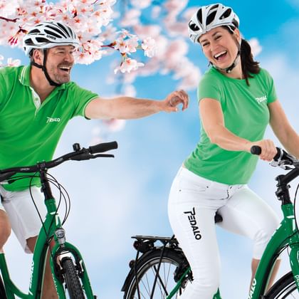 Zwei Radfahrer in grünen Pedalo-Shirts und weißen Helmen fahren grüne Fahrräder unter rosa Kirschblüten vor blauem Himmel.