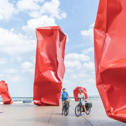 Zwei Radfahrer mit Rädern zwischen großen roten abstrakten Skulpturen auf einer Promenade unter blauem Himmel mit weißen Wolken.