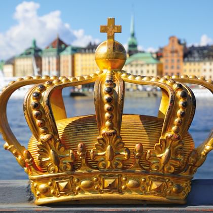 Verzierte goldene Krone mit Kreuz oben im Vordergrund, dahinter Stockholms bunte historische Gebäude und Uferfront über dem Hafen.