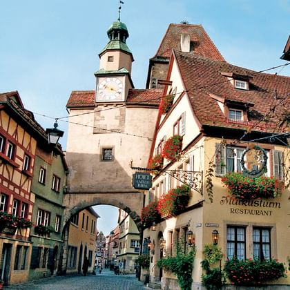 Mittelalterlicher Markusplatz in Rothenburg mit Steinturm-Tor, bunten Fachwerkhäusern und Kopfsteinpflasterstraße unter blauem Himmel.