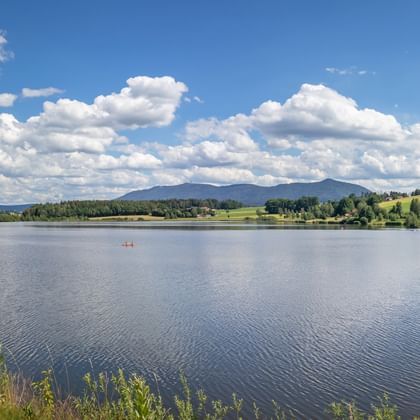 Malerischer See mit ruhigem Wasser in der Region Bayern-Böhmen. Sanfte grüne Hügel und Berge unter blauem Himmel mit weißen Wolken. Kleines Boot auf dem Wasser.
