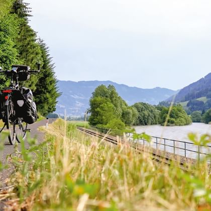 Radfahrer mit Packtaschen auf asphaltiertem Weg entlang der Mur, umgeben von grünen Wiesen, Bäumen und Bergen im Hintergrund.