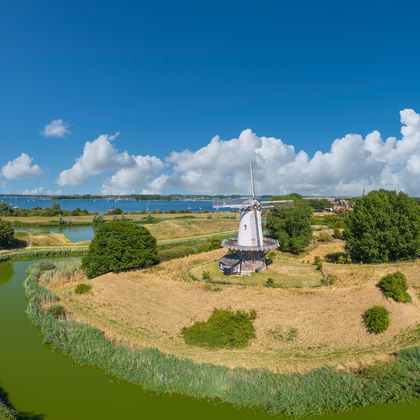 Luftaufnahme einer weißen Windmühle auf einer Halbinsel, umgeben von grünen Wasserläufen in Zeeland. Blauer Himmel mit weißen Wolken.