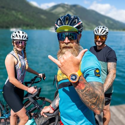Drei Gravelbiker mit Helmen und Sonnenbrillen auf einem Holzsteg an einem türkisfarbenen Alpensee in Kärnten, Berge im Hintergrund.