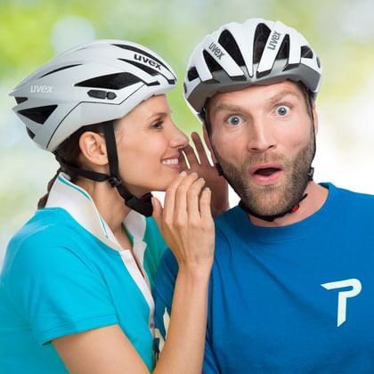 Frau in türkisfarbenem Shirt und weißem Helm flüstert überraschtem Mann in blauem Shirt zu. Beide tragen weiße Fahrradhelme vor unscharfem Hintergrund.
