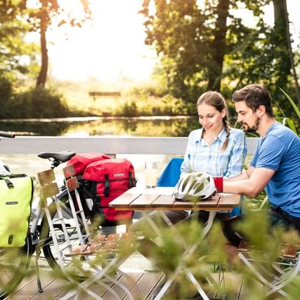 Zwei Radfahrer sitzen an einem Holz-Picknicktisch mit Tourenrädern und Packtaschen in der Nähe, umgeben von üppigem Grün in Thüringen.