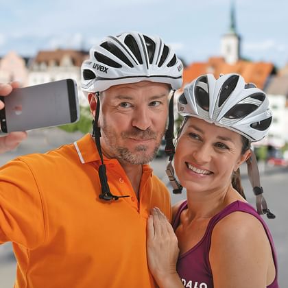Zwei lächelnde Radfahrer mit weißen Helmen machen ein Selfie. Mann im orangenen Shirt und Frau im lila Top posieren vor historischem Stadtplatz.