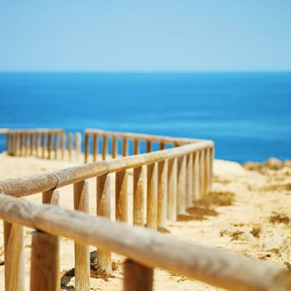 Holzgeländer entlang sandiger Klippenkante an der Algarve mit blauem Meer und klarem Himmel. Ein kleines Segelboot am Horizont.