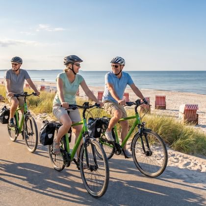 Vier Radfahrer mit Helmen und Packtaschen auf grünen Rädern auf einem Küstenweg. Sandstrand mit Strandkörben und ruhiger See im Hintergrund.