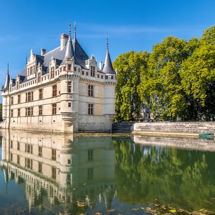 Weißes Renaissance-Schloss mit spitzen Türmchen spiegelt sich im umgebenden Wassergraben, eingerahmt von grünen Bäumen unter blauem Himmel.