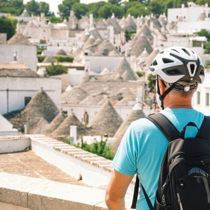 Rückansicht eines Radfahrers mit weißem Helm und blauem Shirt mit Blick auf traditionelle Trulli-Häuser mit Kegeldächern in Apulien.