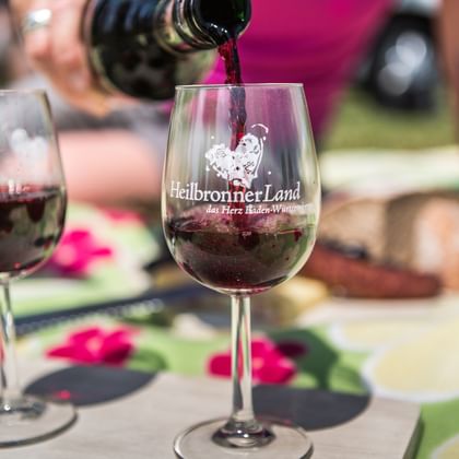 Rotwein wird in ein Glas mit Heilbronner Land Logo eingeschenkt. Ein weiteres gefülltes Weinglas und Speisen sind im Hintergrund sichtbar.