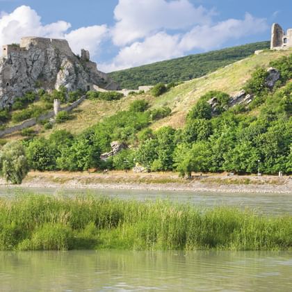 Two medieval castle ruins perched on rocky hills overlooking the Danube River, with green vegetation and reeds along the riverbank.