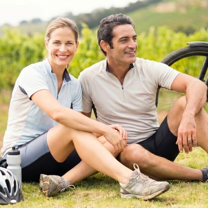 Paar sitzt auf Wiese in Weinberg mit Fahrrad und Helmen. Weinreben und sanfte Hügel im Hintergrund sichtbar.