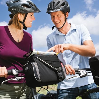 Two cyclists with helmets standing with their bikes equipped with handlebar bags, smiling at each other under a blue sky with clouds.