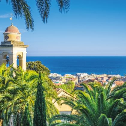 Glockenturm mit Kuppel, umgeben von Palmen, mit Blick auf die Küstenstadt San Remo und das Mittelmeer unter blauem Himmel.