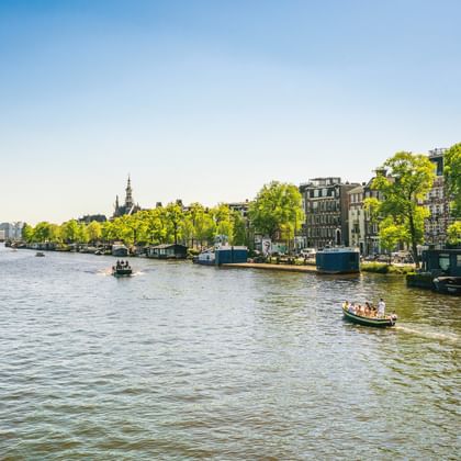 Breite Gracht in Amsterdam mit Hausbooten an baumgesäumten Ufern, historischen Gebäuden und modernen Türmen in der Ferne unter blauem Himmel.