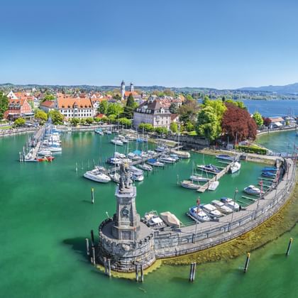 Luftaufnahme des Lindauer Hafens am Bodensee mit Leuchtturm, Jachthafen, historischen Gebäuden mit roten Dächern und türkisfarbenem Wasser.