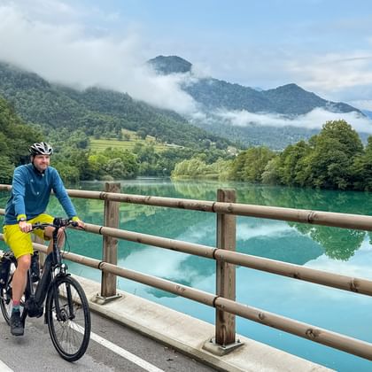 Radfahrer in blauer Jacke und gelber Hose auf einer Brücke mit Holzgeländer über einem türkisfarbenen See in Slowenien mit nebligen Bergen im Hintergrund.