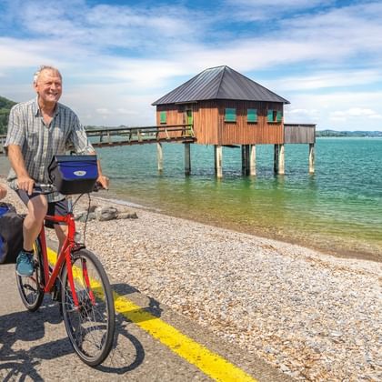 Zwei Radfahrer mit roten Rädern und blauen Taschen stehen am Kiesstrand des Bodensees. Ein hölzernes Pfahlhaus steht im türkisfarbenen Wasser.