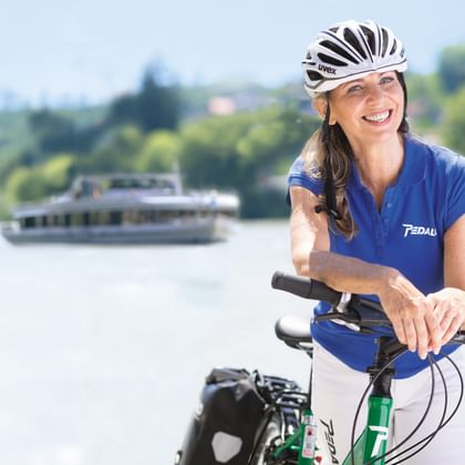 Lächelnde Frau mit Helm auf Fahrrad am Fluss. Ausflugsschiff und grüne Landschaft im Hintergrund unter blauem Himmel sichtbar.