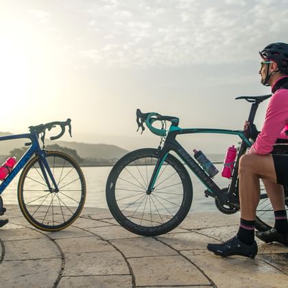 Radfahrer rasten am Wasser in Dalmatien Zwei Radfahrer in rosa-schwarzer Radkleidung mit Rennrädern ruhen auf Steinpflaster neben ruhigem Wasser zur goldenen Stunde.