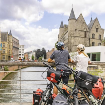 Zwei Radfahrer mit Tourenrädern und Packtaschen pausieren auf einer Brücke über einem Kanal in Flandern, mittelalterliche Steingebäude sichtbar.