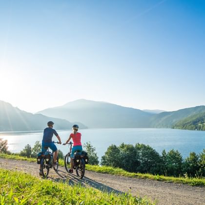 Two cyclists on a gravel path beside a Carinthian lake, surrounded by green hills and mountains under a clear blue sky with morning sunlight.