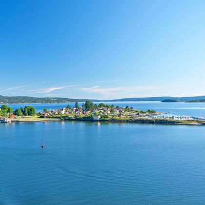 Aerial view of a small island in Oslofjord with residential houses, trees, and a dock. Blue water surrounds the island with hills visible.