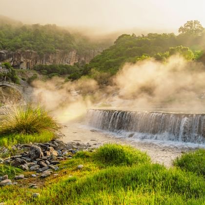 Historische Steinbogenbrücke und Wasserfall in nebelverhangener albanischer Landschaft mit grünen Hügeln, fließendem Fluss und Morgenlicht.