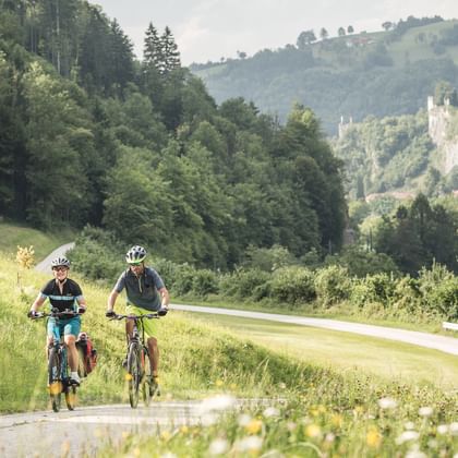 Zwei Radfahrer auf einem asphaltierten Weg durch grüne Wiesen entlang des Ennsradwegs mit bewaldeten Hügeln im Hintergrund.