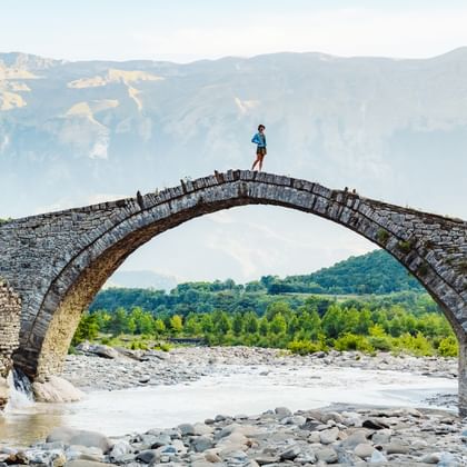 Person auf einer alten Steinbogenbrücke über einem steinigen Flussbett in Albanien, mit Bergen und grünen Tälern im Hintergrund.