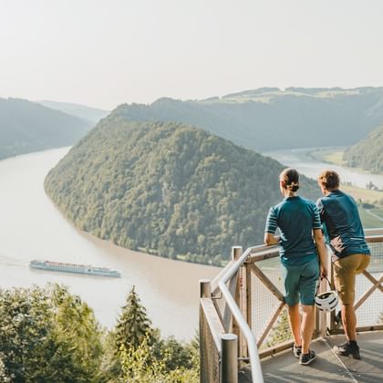 Zwei Radfahrer auf einer Aussichtsplattform mit Blick auf die Donau, die sich durch bewaldete Hügel schlängelt. Ein Frachtschiff ist auf dem Fluss sichtbar.