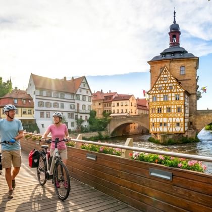Zwei Radfahrer schieben ihre Räder auf einer Holzbrücke in Bamberg, mit dem historischen Fachwerkrathaus am Fluss im Hintergrund.