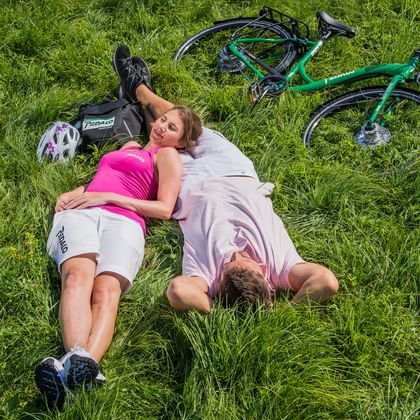 Two cyclists lying on green grass with a turquoise bicycle beside them. They are wearing cycling gear and helmets are visible nearby.