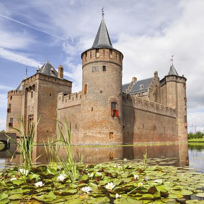 Medieval brick castle with round towers and spire surrounded by moat with water lilies under blue sky with clouds.