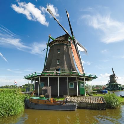 Dunkelgrüne Windmühle mit orangen Flügeln an Wasserweg in Noord-Holland. Blauer Himmel mit weißen Wolken, grünes Schilf, weitere Mühlen sichtbar.