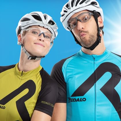 Zwei Radfahrer mit weißen Helmen und Brillen posieren vor blauem Himmel. Einer trägt ein gelbes, der andere ein türkisfarbenes Trikot.