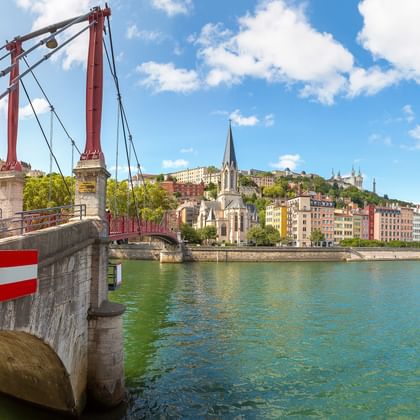Rote Hängebrücke über die Rhône in Lyon mit bunten Gebäuden und Kirchturm entlang des Ufers unter blauem Himmel.