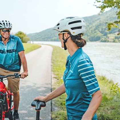 Two cyclists in turquoise jerseys on a paved path along the Danube River, with green hills and water in the background.