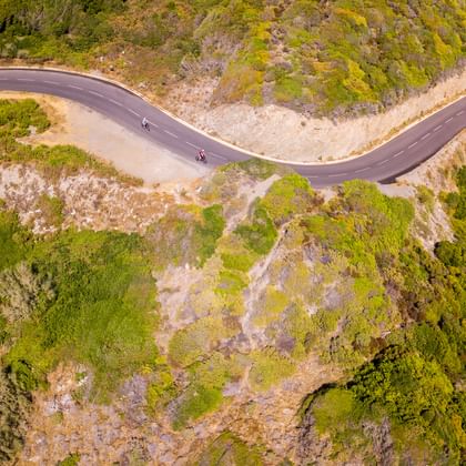 Luftaufnahme von drei Radfahrern auf einer kurvenreichen Bergstraße auf Korsika, umgeben von grüner mediterraner Vegetation und felsigem Gelände.