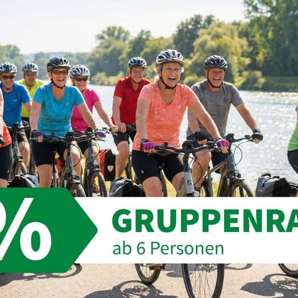 Gruppe von Radfahrern mit Helmen und bunten Shirts auf einem asphaltierten Weg neben einem Fluss mit Bäumen im Hintergrund.